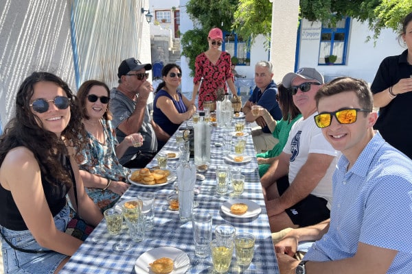 Friends dining al fresco at a long table with food and drinks, backed by white buildings and greenery.